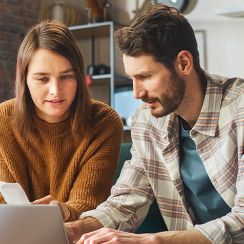 A woman and a man sit closely together, focused on a laptop. They appear to be discussing or analyzing information, set against a cozy, modern room with plants and bookshelves.