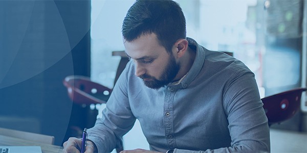 A man in a light blue shirt is writing with a pen on a piece of paper while sitting at a table in a well-lit café environment, surrounded by modern furniture.
