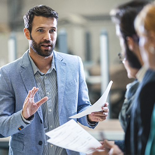A man in a light blue blazer is gesturing while holding papers, engaged in discussion with two people in an indoor setting, likely a workplace or meeting space.
