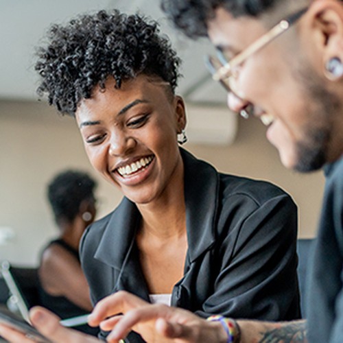 A woman and a man are smiling and engaging with a tablet. They appear to be sharing an enjoyable moment in a modern indoor setting, with soft lighting and minimalistic decor in the background.
