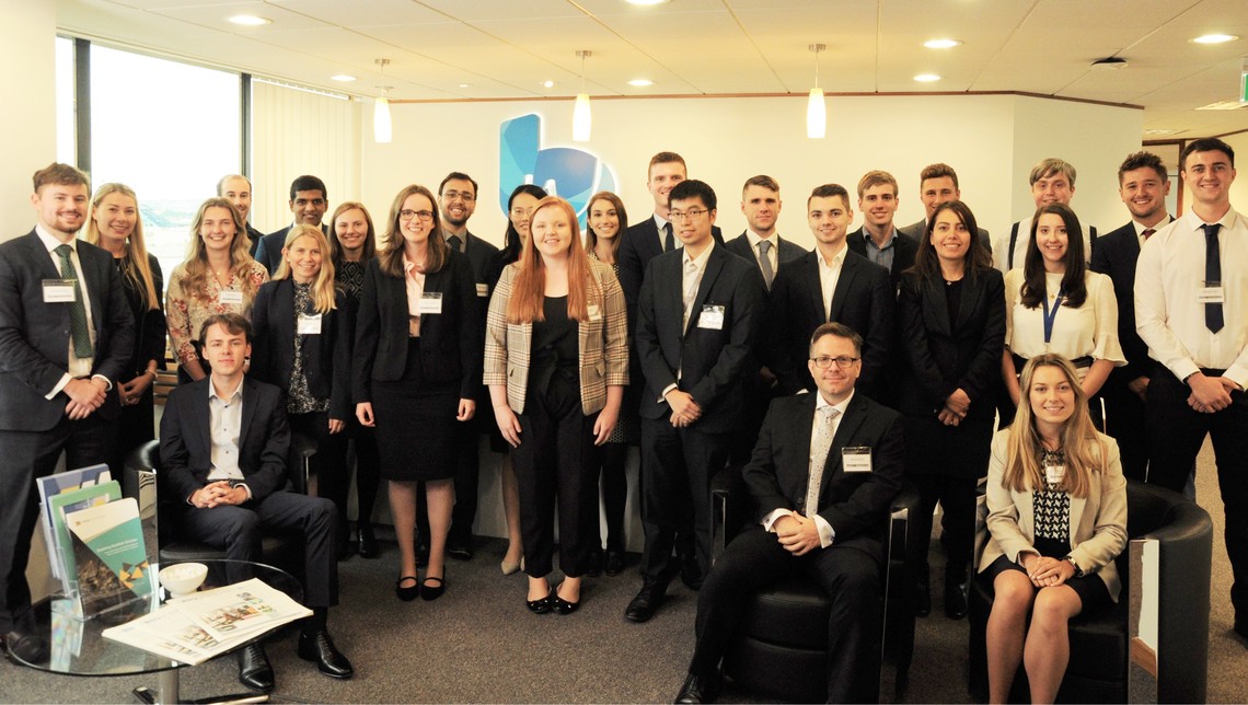 A group of young professionals poses together in a well-lit office environment, smiling and dressed in business attire, with a logo on the wall behind them and printed materials on a table nearby.