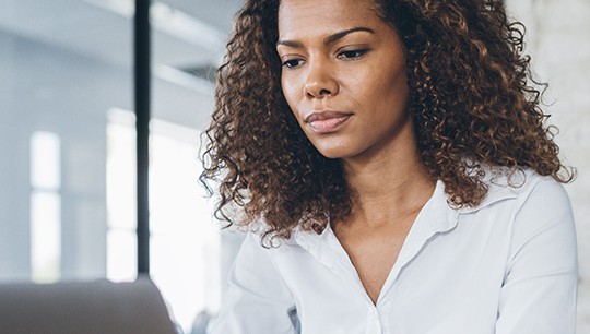 A woman with curly hair sits at a desk, focused on her laptop. Bright natural light filters through large windows in a modern office space with a clean, minimalistic design.