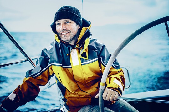 A man in a bright yellow and black waterproof jacket and a beanie is steering a boat, smiling, with calm blue waters and a cloudy sky in the background.