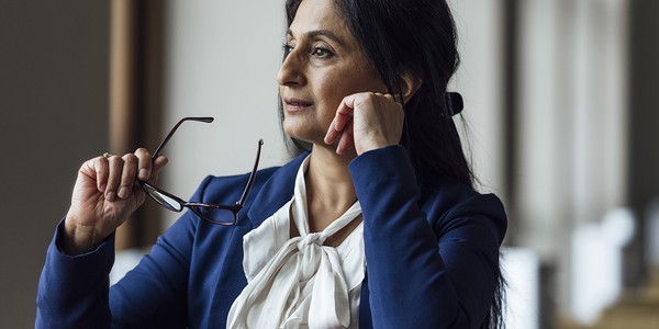 A woman in a blue blazer rests her chin on her hand, holding eyeglasses with the other hand, gazing thoughtfully. Soft light filters in from a nearby window, creating a contemplative atmosphere.