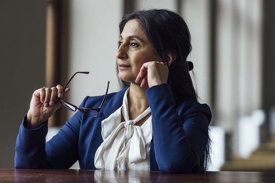 A woman in a blue blazer rests her chin on her hand, holding eyeglasses with the other hand, gazing thoughtfully. Soft light filters in from a nearby window, creating a contemplative atmosphere.