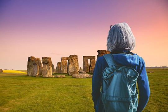 A person with gray hair, wearing a blue hoodie and a backpack, stands facing a stone monument at sunset. Vibrant hues fill the sky, with fields visible in the background.