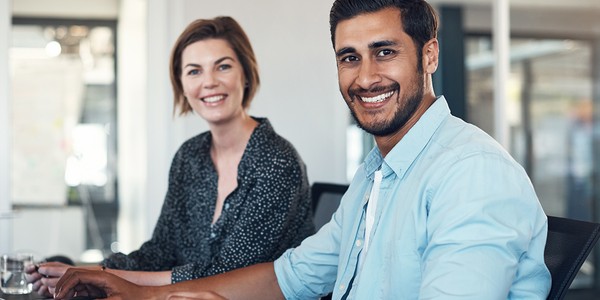 Two colleagues, a man and a woman, smile while sitting at a desk, with computers in front of them. The setting is a modern office, featuring glass walls and natural lighting.