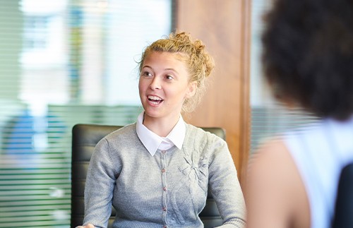 A young woman with curly hair, wearing a gray cardigan and white collared shirt, is smiling and speaking in a modern office setting with glass partitions in the background.