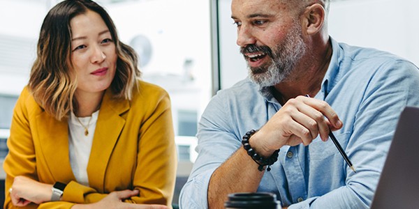 A woman and a man are engaged in conversation at a table. The man, holding a pen, is animated, while the woman listens attentively. A coffee cup and papers are on the table, with a modern office backdrop.