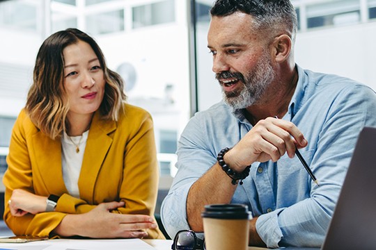 A woman and a man are engaged in conversation at a table. The man, holding a pen, is animated, while the woman listens attentively. A coffee cup and papers are on the table, with a modern office backdrop.