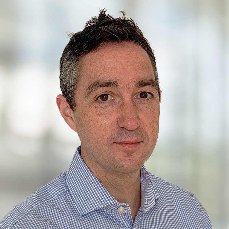 A man with short, dark hair and a light blue checkered shirt faces the camera, presenting a neutral expression. He is set against a blurred, soft-focus background.