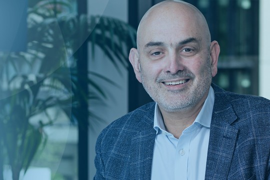 A man with a light beard smiles while seated in an office environment. He wears a blue patterned blazer and a white shirt, with greenery visible in the background.