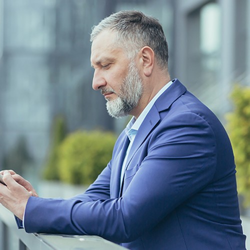 A man in a blue suit is focused on his smartphone while leaning against a railing. He's outdoors, surrounded by modern buildings and greenery, suggesting a professional atmosphere.