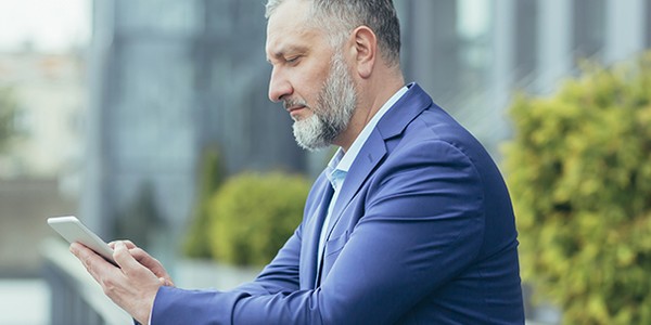 A man in a blue suit is focused on his smartphone while leaning against a railing. He's outdoors, surrounded by modern buildings and greenery, suggesting a professional atmosphere.