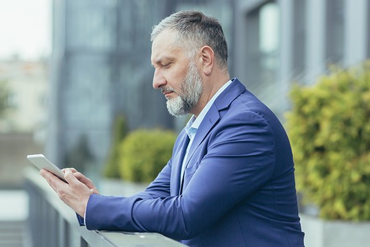 A man in a blue suit is focused on his smartphone while leaning against a railing. He's outdoors, surrounded by modern buildings and greenery, suggesting a professional atmosphere.
