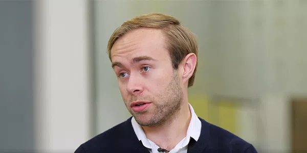 A man with light brown hair is speaking, wearing a dark sweater over a white shirt. The background is blurred, suggesting an indoor setting with light colors.