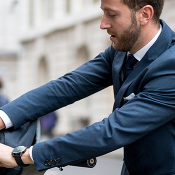A man in a suit is locking a bicycle at a bike rack on a city street. Pedestrians and buildings are visible in the background, indicating a bustling urban environment.