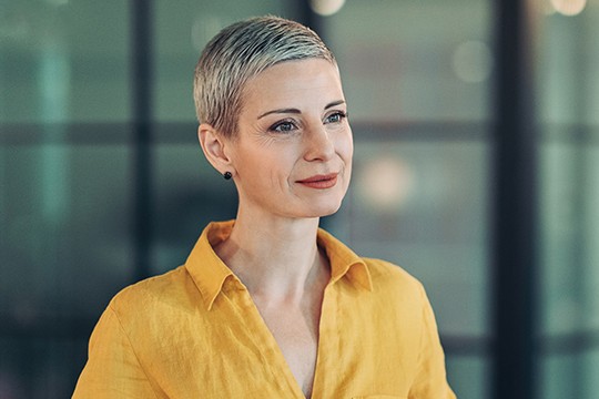 A woman with short, light hair wears a bright yellow shirt. She appears thoughtful, looking slightly to the side, surrounded by a modern, softly lit environment with blurred background elements.