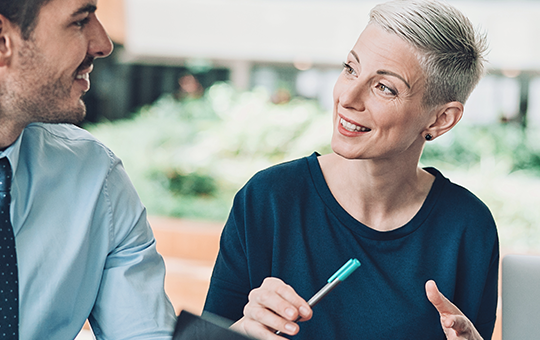 A group of four professionals engage in lively discussion, with one woman smiling and gesturing. They are seated around a table in a bright, plant-filled office environment.