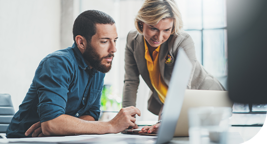 A man and woman examine a laptop screen together, with the woman leaning slightly over the man's shoulder. They are in a well-lit, modern office setting, with greenery visible outside.