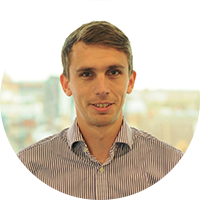 A man with short brown hair and a slight smile stands facing the camera, wearing a striped shirt. A cityscape is blurred in the background, suggesting an office environment.