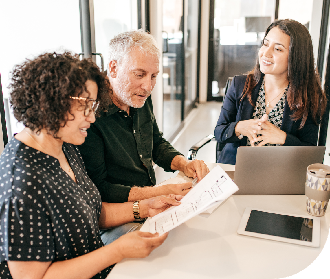 Three people are engaged in a discussion around a table. One person is reviewing documents, while another gestures animatedly. A laptop and a coffee cup are present in a modern office setting.