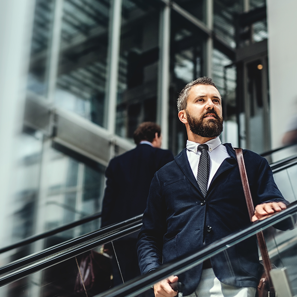 A businessman, dressed in a suit, confidently ascends an escalator. Surrounding him are blurred figures of other professionals in a modern, glass-filled building, suggesting a busy urban environment.