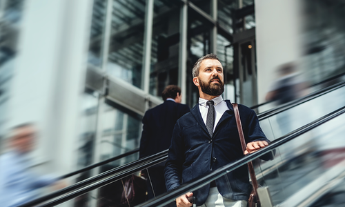 A businessman, dressed in a suit, confidently ascends an escalator. Surrounding him are blurred figures of other professionals in a modern, glass-filled building, suggesting a busy urban environment.