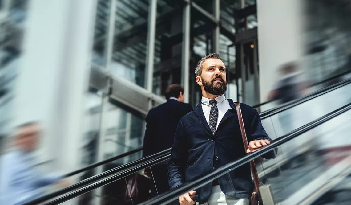 A businessman, dressed in a suit, confidently ascends an escalator. Surrounding him are blurred figures of other professionals in a modern, glass-filled building, suggesting a busy urban environment.