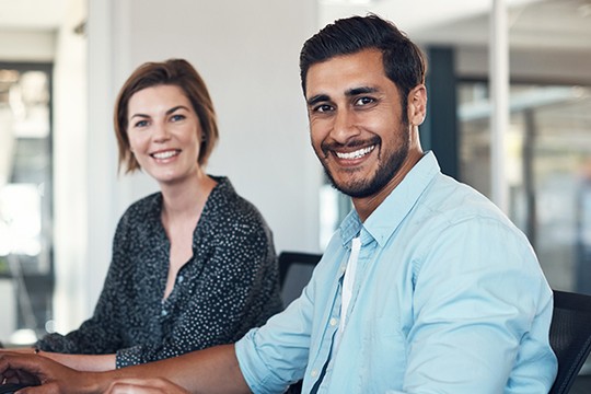 Two people sit at a table in a bright office setting. They smile at the camera; the woman wears a patterned blouse, while the man is in a light blue shirt.