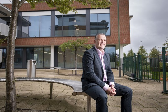 A man in a suit sits on a curved metal bench, smiling. Behind him is a modern building with large glass windows and a landscaped area, creating a professional outdoor setting.