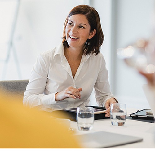 A woman in a white shirt is smiling and engaging in conversation during a meeting. She sits at a table with clear water glasses, surrounded by colleagues in a bright, modern office setting.