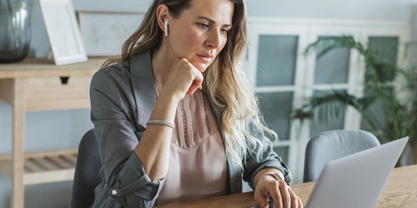 A woman with long hair, wearing a gray blazer and seated at a wooden desk, attentively types on a laptop, surrounded by a bright, minimalistic home office setting with greenery.
