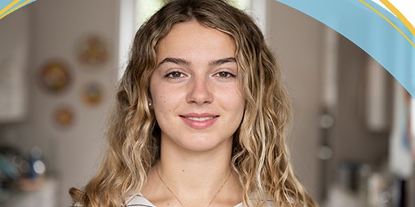 Female with wavy hair smiles at the camera. She is indoors, with a kitchen-like background featuring decorative plates on the wall, conveying a warm, inviting atmosphere.