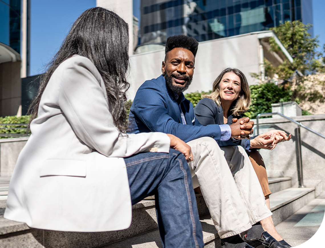 Three professionals sit on stone steps, engaged in conversation. A man in a blue shirt smiles between two women, one in a light jacket and the other in a gray blazer, surrounded by urban buildings and greenery.