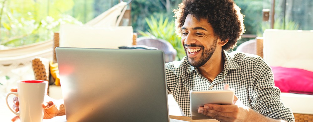 A man with curly hair is smiling while holding a tablet in one hand and a coffee cup in the other, working on a laptop in a cozy, well-lit indoor setting surrounded by greenery.