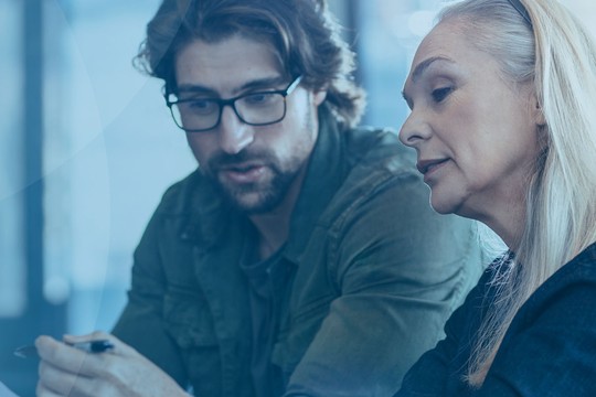 Two individuals, a man and a woman, are engaged in conversation while examining a document. They are seated in a well-lit indoor environment with a blue background.