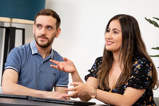 Two individuals are engaged in conversation at a table. A woman gestures expressively while speaking, and a man listens attentively, both appearing to discuss a topic in a bright, modern room.