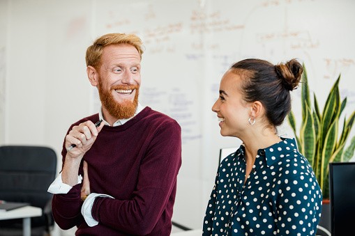 Two people are engaged in a cheerful conversation. The man, with a red beard, smiles while holding a pen. They are in a bright office space with plants and whiteboard notes in the background.