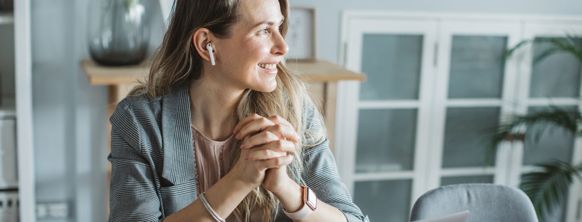 A woman with wireless earbuds smiles while clasping her hands, seated at a table in a bright, modern room with plants and glass-paneled cabinets in the background.