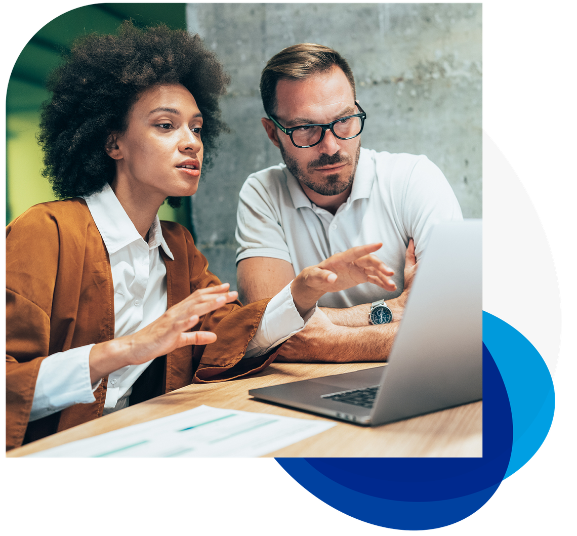 A woman with curly hair gestures animatedly while discussing with a man in glasses. They sit at a wooden table, focused on a laptop screen, in a modern, well-lit workspace.