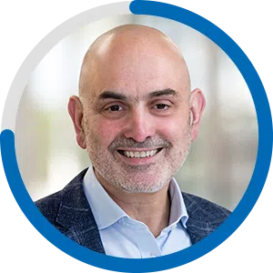 A man with a shaved head and a well-groomed beard smiles warmly. He wears a light blue shirt under a dark jacket. The background is softly blurred, suggesting an indoor setting.