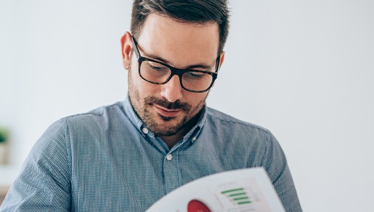 A man in a blue checkered shirt and glasses sits at a desk, intently reading a document showing graphs and charts in a bright, minimalistic office environment.
