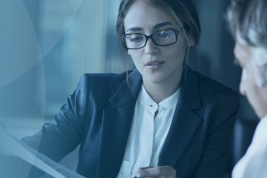 A professional-looking woman in glasses reviews a document, engaged in discussion with a man. They sit in a well-lit office with large windows, suggesting a business context.