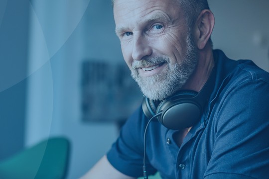 A smiling man wearing headphones is sitting at a desk, reviewing papers. The background includes a lamp and wall art, creating a cozy work environment.