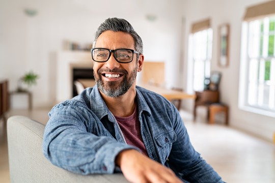 A smiling man wearing glasses and a blue shirt sits on a couch, extending his arm outward. He is in a well-lit, modern living room with furniture and plants in the background.