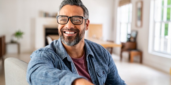 A smiling man wearing glasses and a blue shirt sits on a couch, extending his arm outward. He is in a well-lit, modern living room with furniture and plants in the background.