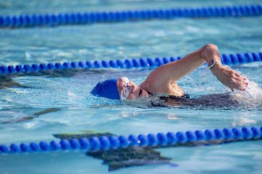 A swimmer in a blue cap and goggles performs freestyle strokes in a pool, splashing water as they move. Blue lane markers line the pool's edge under a bright, clear sky.