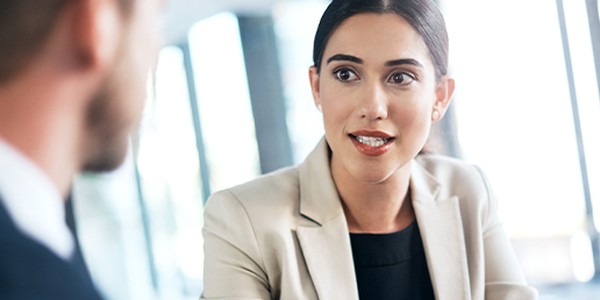 A professional woman, dressed in a light-colored blazer, is engaged in conversation, actively listening and responding, while seated across from a man in a dark suit in a bright, modern office setting.