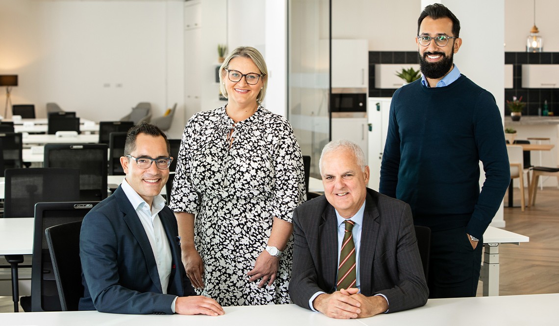 Four professionals pose for a group photo in a modern office setting. Two sit at a table, while two stand behind them, all smiling. The background features desks and a kitchenette.